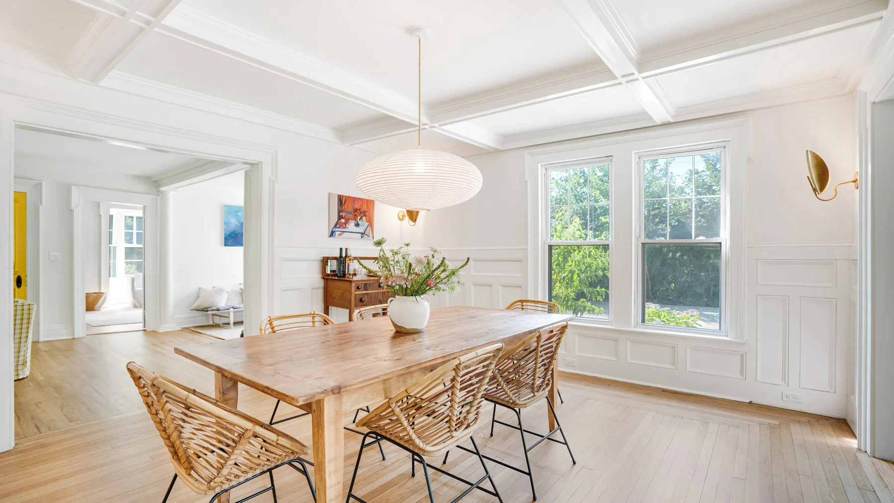 A bright dining room with a wooden table and woven seating beside large windows.
