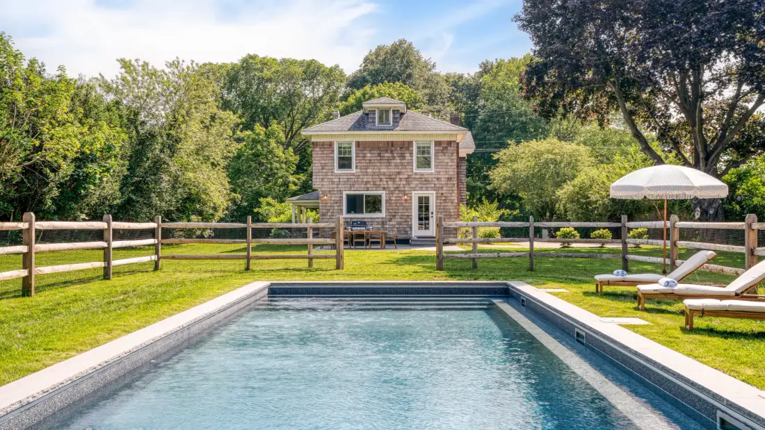 An outdoor swimming pool sits in front of a small brick building and fenced lawn.