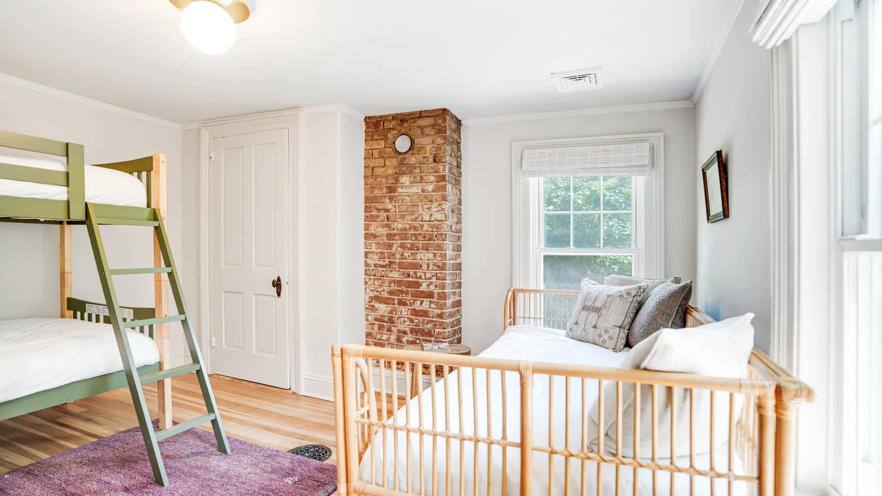 A bright bedroom with a wooden bunk bed, crib, and exposed brick chimney.
