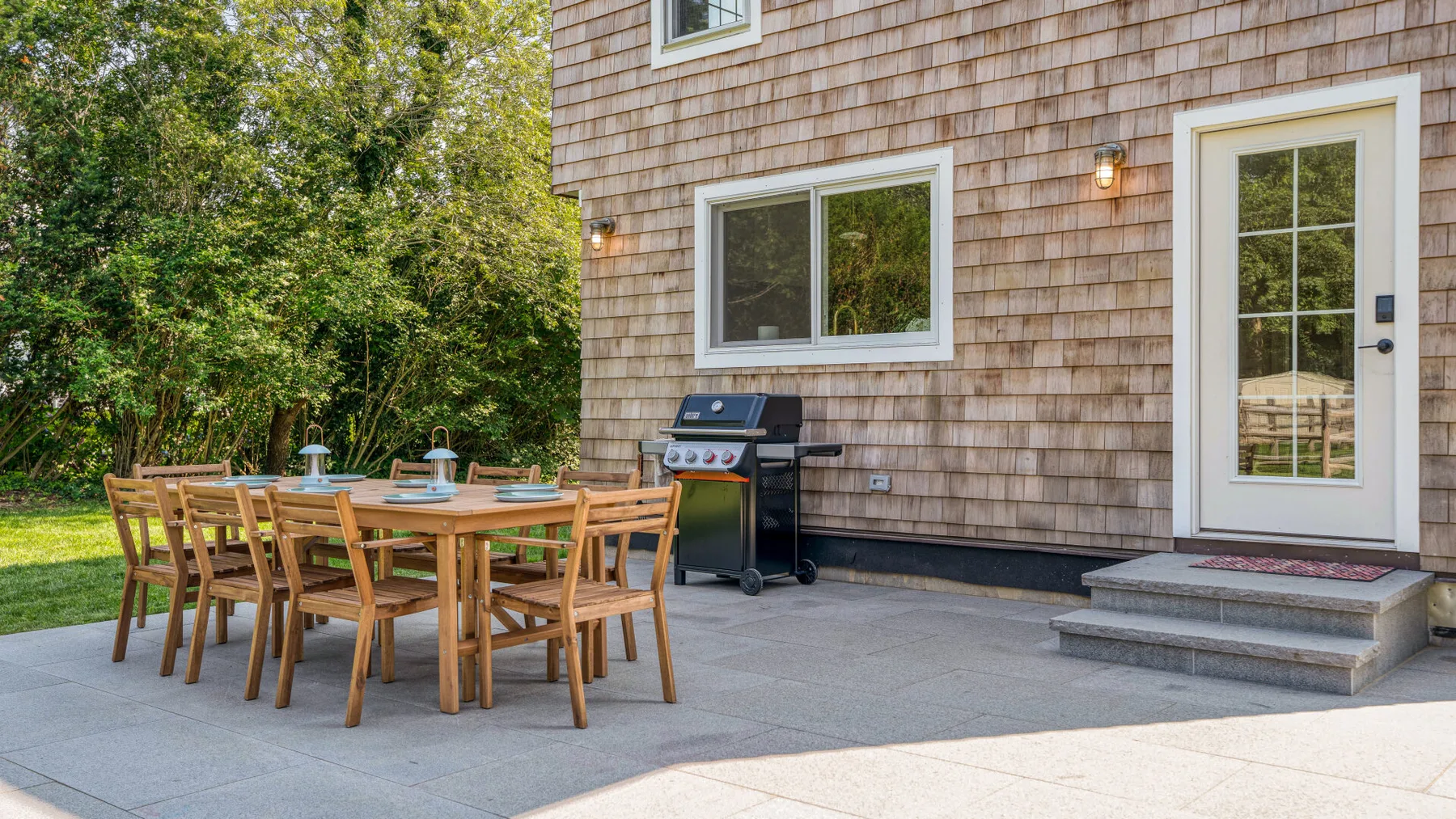 An outdoor patio with a wooden dining table and grill beside a house.