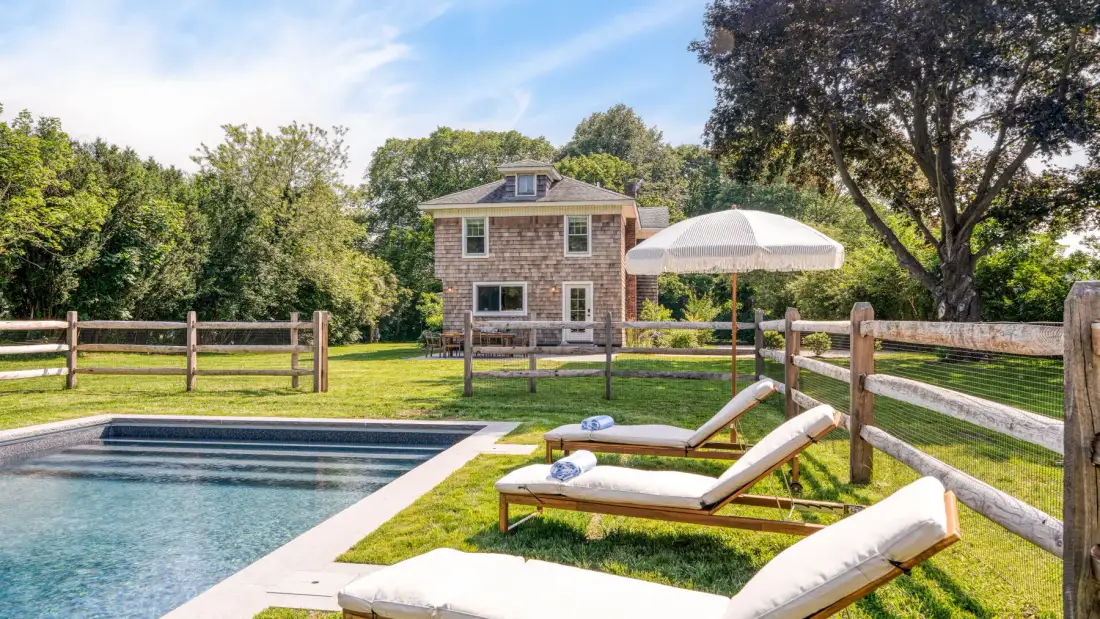 A backyard pool and lounge chairs sit beside a fenced lawn and brick house.
