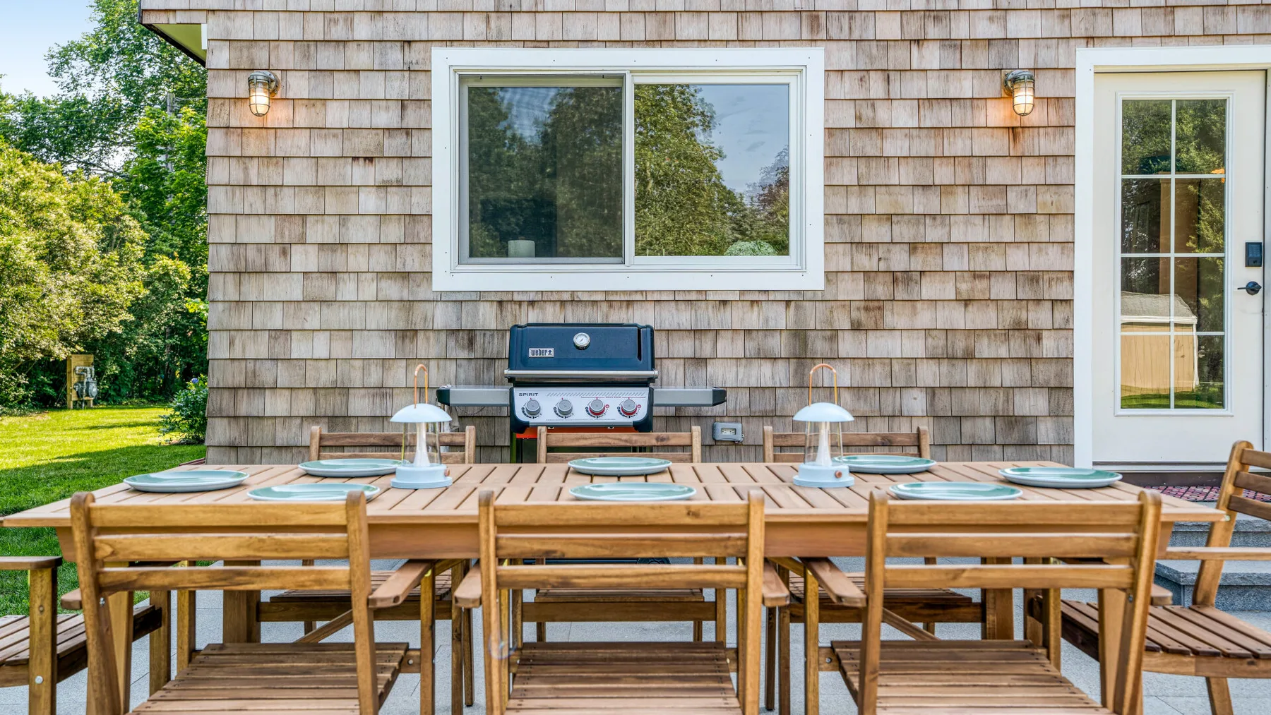 An outdoor dining table with wooden seating stands beside a shingled house.