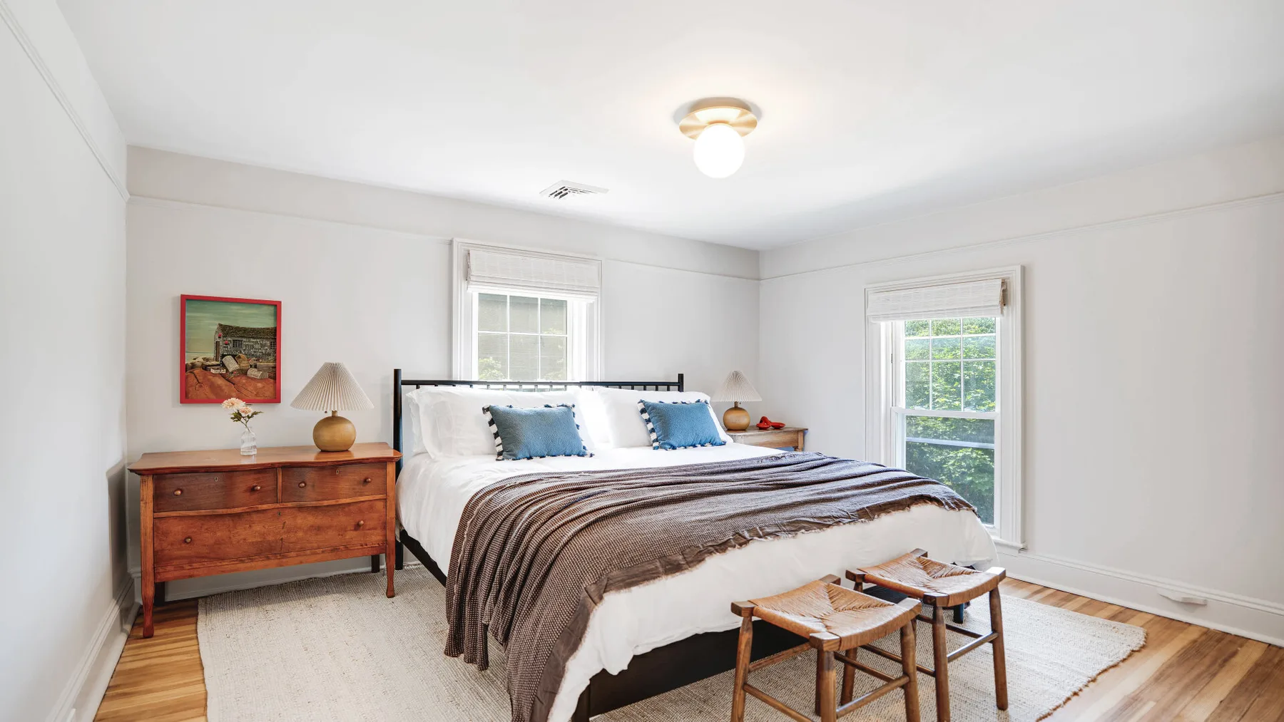 A bright bedroom with a large bed, wooden dresser, and two windows.