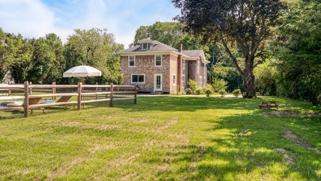 A brick house stands beyond a broad lawn, wooden fence, and shade trees.