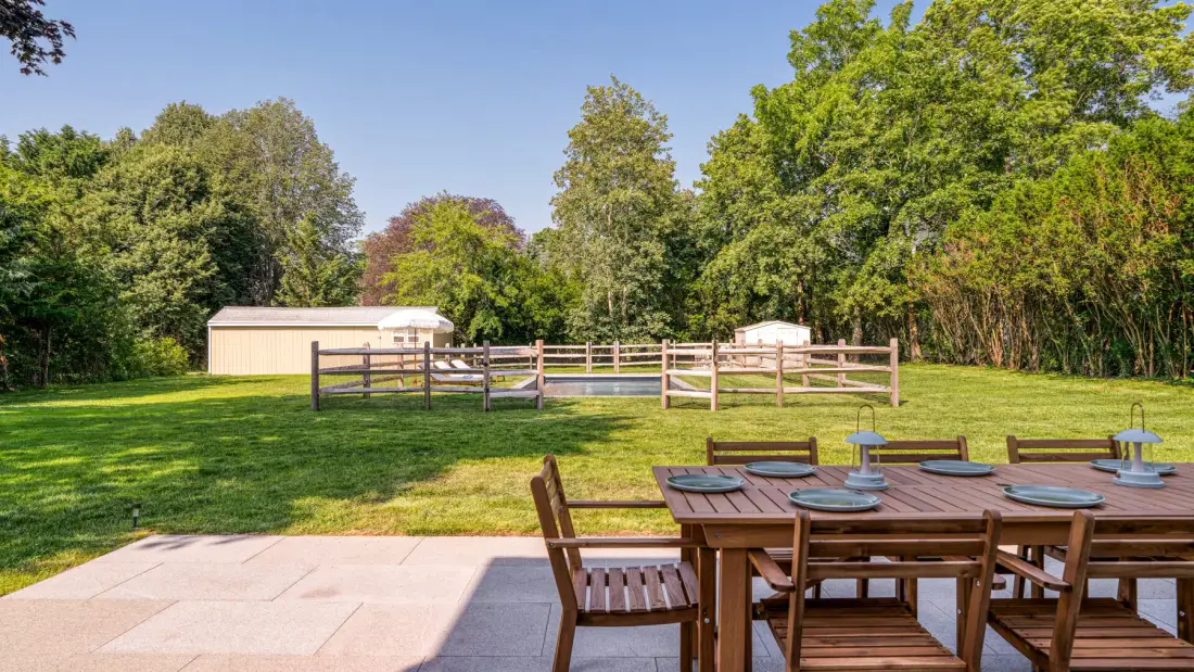 An outdoor dining area overlooks a grassy yard enclosed by trees and fencing.