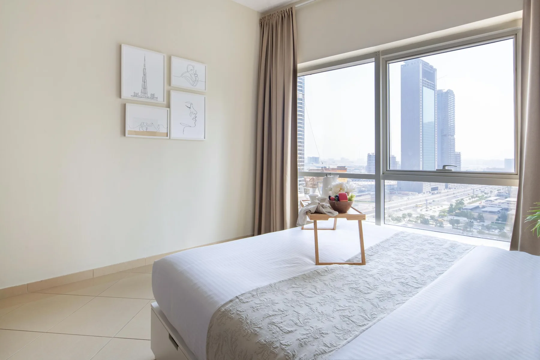 Un dormitorio con una cama junto a grandes ventanas con vistas a edificios cercanos.