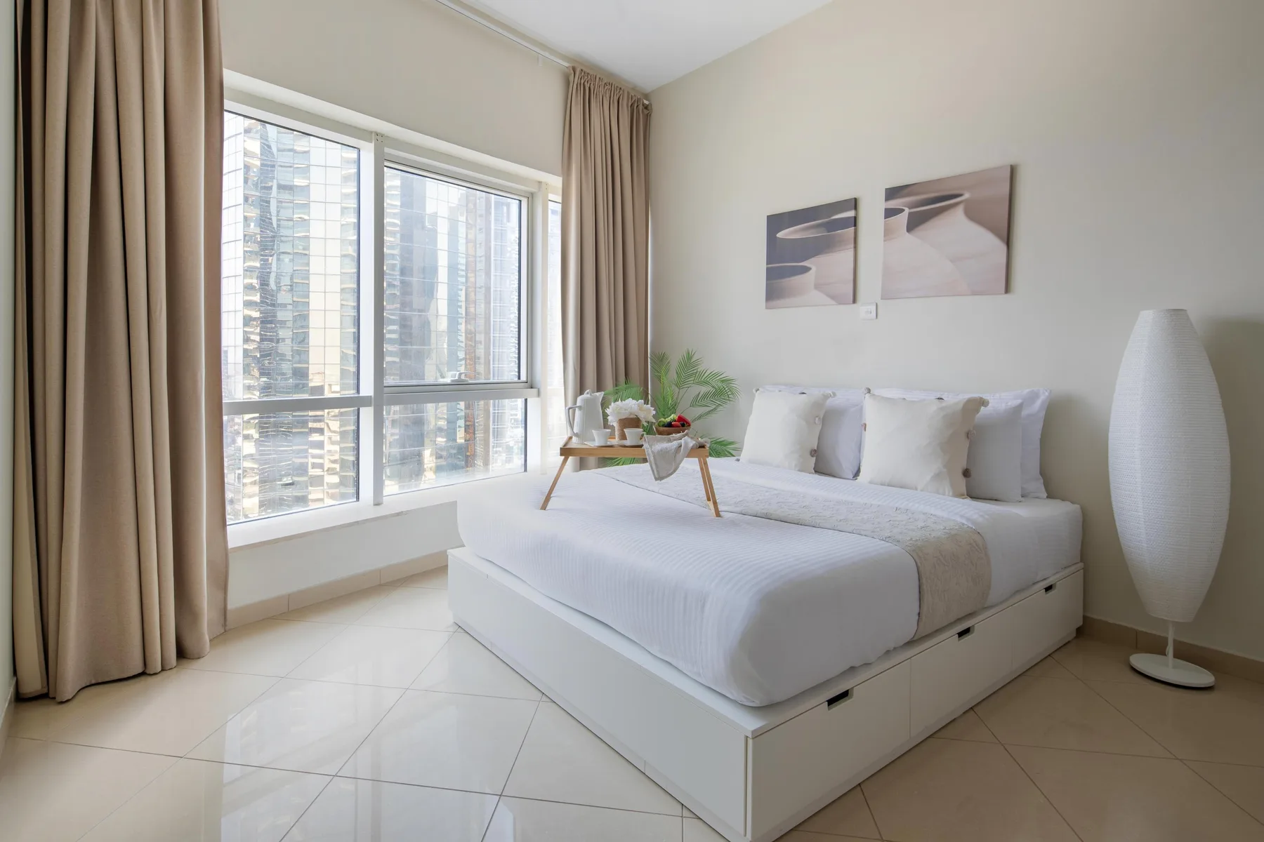 Un dormitorio luminoso con una cama blanca junto a grandes ventanas y cortinas beige.