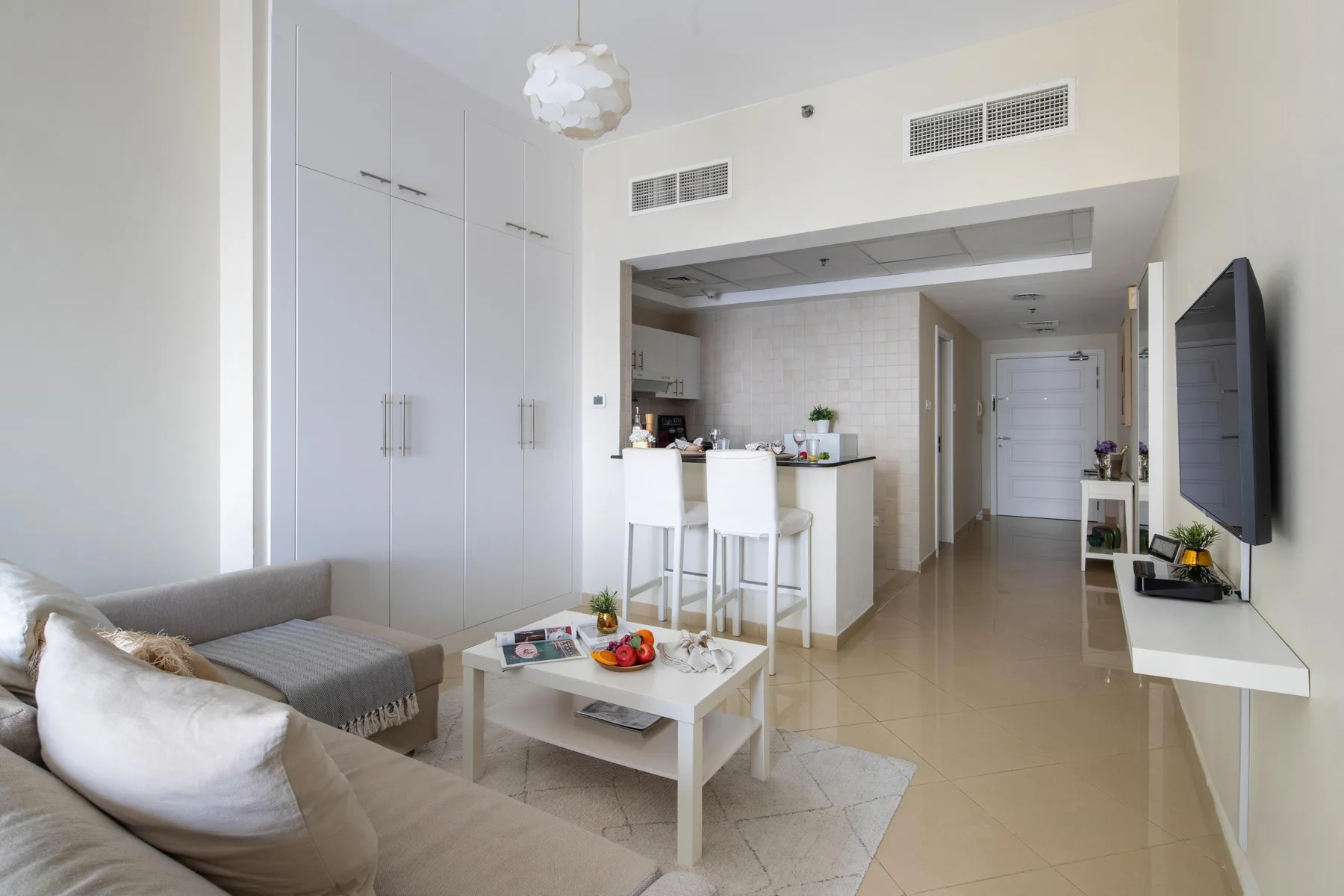 Una luminosa sala de estar se abre a una cocina compacta, con asientos y un televisor en la pared.