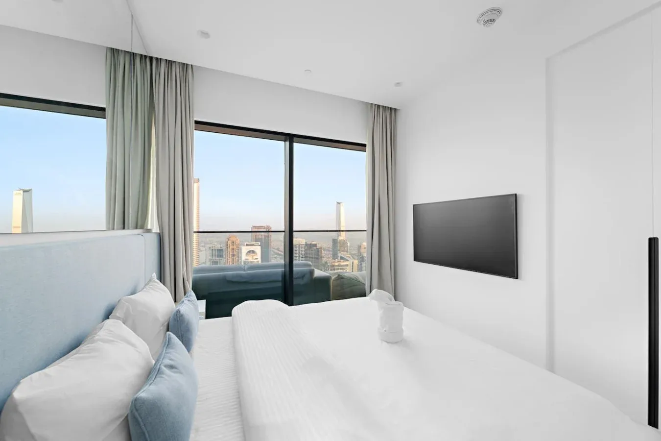 Un dormitorio luminoso con una cama frente a un televisor de pared y ventanas grandes.