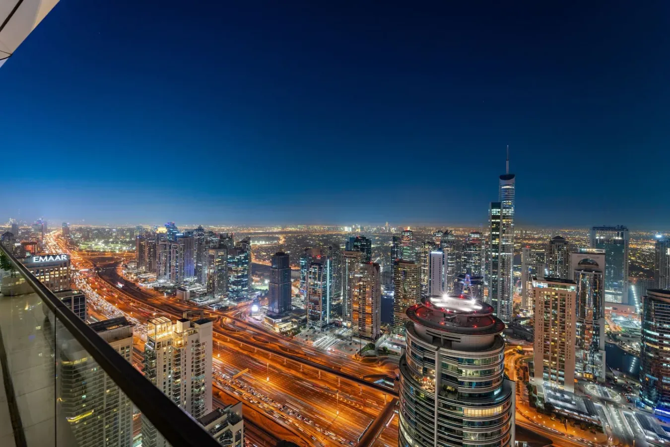 Panorámica nocturna de la ciudad desde un balcón, con carreteras iluminadas y edificios altos.