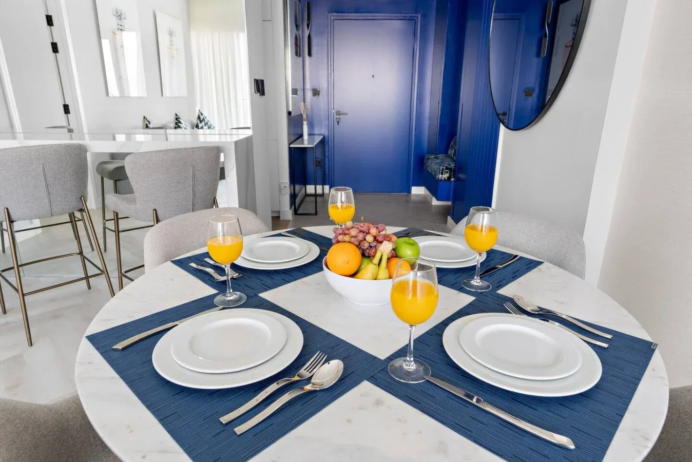 Una mesa de comedor está puesta con platos, manteles individuales azules, fruta y zumo de naranja.