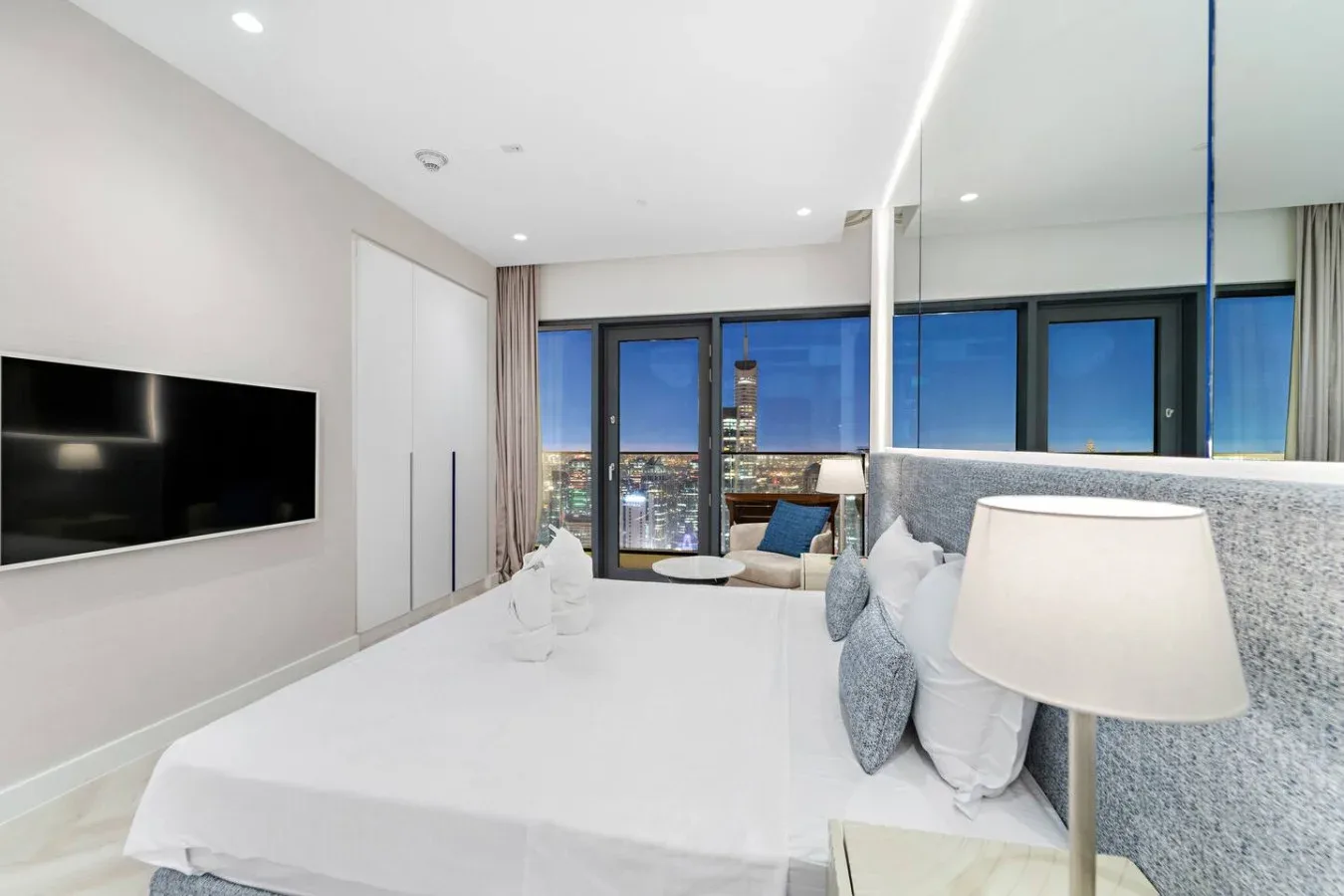 Un dormitorio luminoso con una cama grande, un televisor en la pared y vistas de la ciudad por las ventanas.