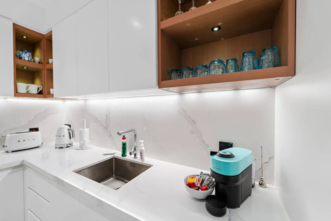 Una luminosa encimera de cocina con fregadero, pequeños electrodomésticos y estantes abiertos.