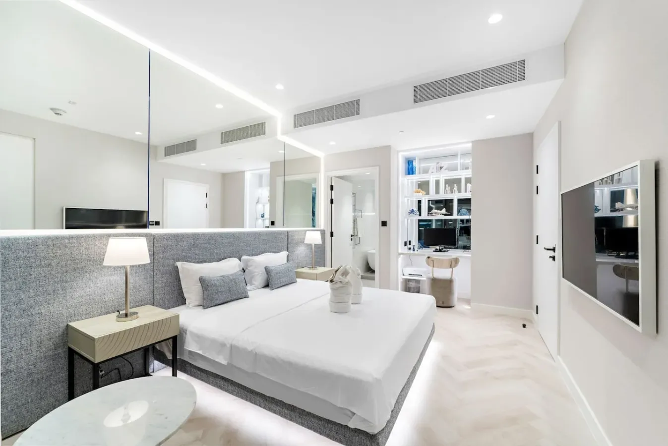 Un dormitorio luminoso tiene una cama bien tendida junto a un cabecero gris tapizado.