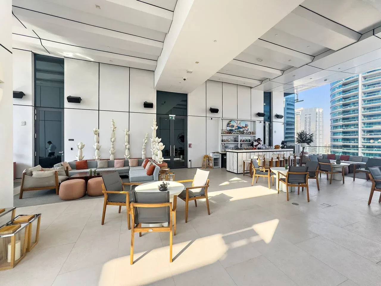 Un salón interior luminoso con asientos alrededor de mesas y grandes ventanales.