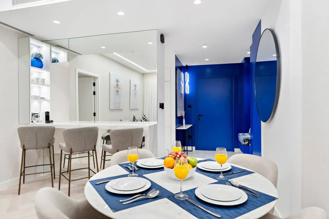 Un comedor con la mesa puesta está junto a una cocina luminosa y una entrada azul al baño.