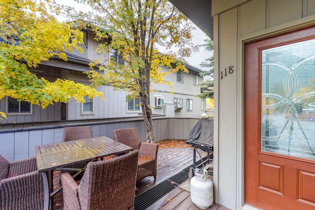 A wooden outdoor deck has a dining table and seating beside a glass-paneled door.