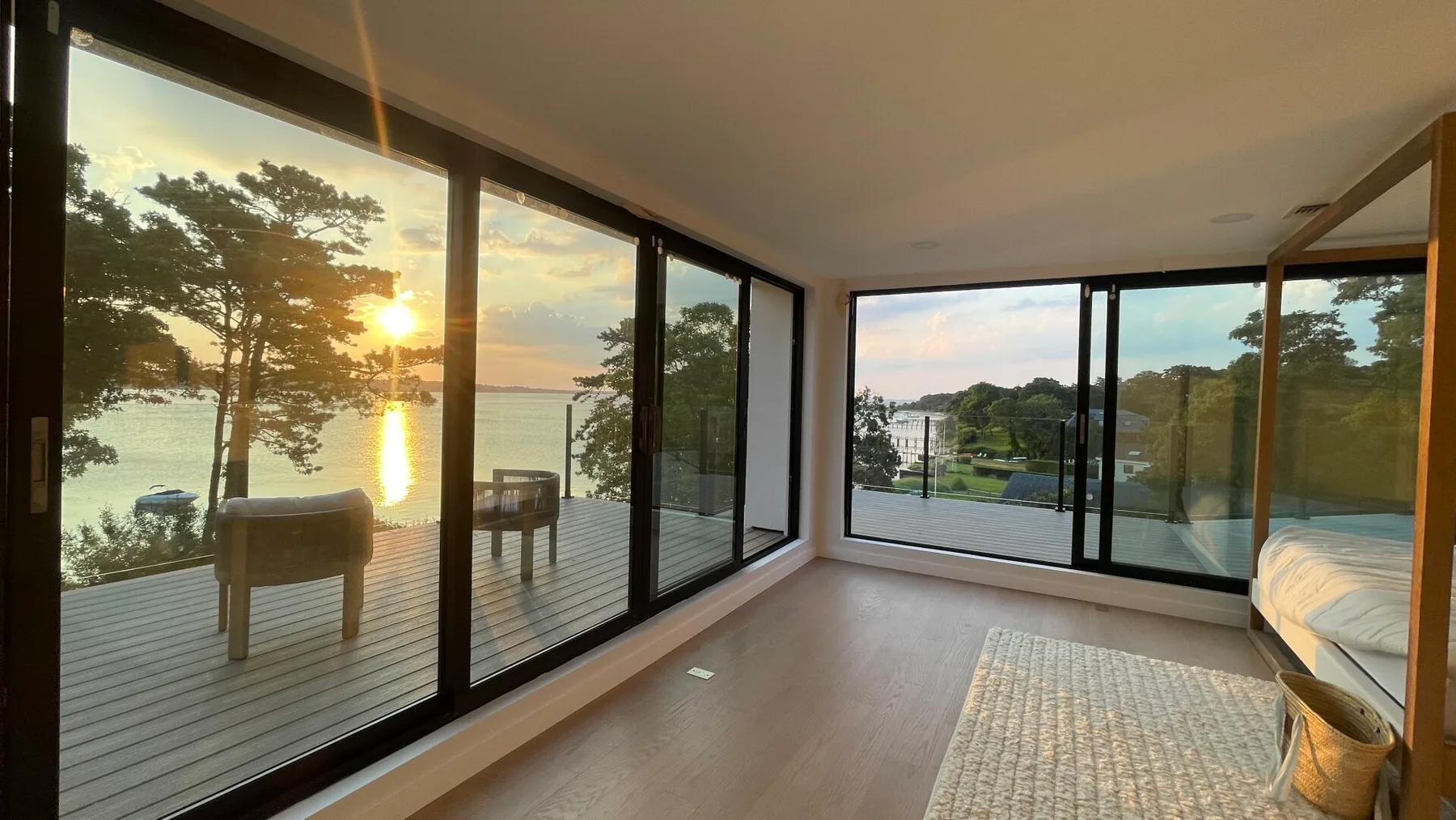 A glass-walled bedroom opens onto a deck overlooking the water at sunset.