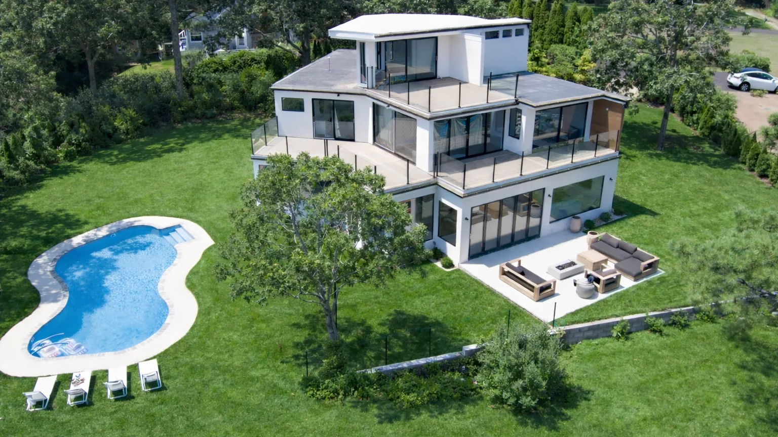 A modern multi-level house sits beside a curved swimming pool on a green lawn.