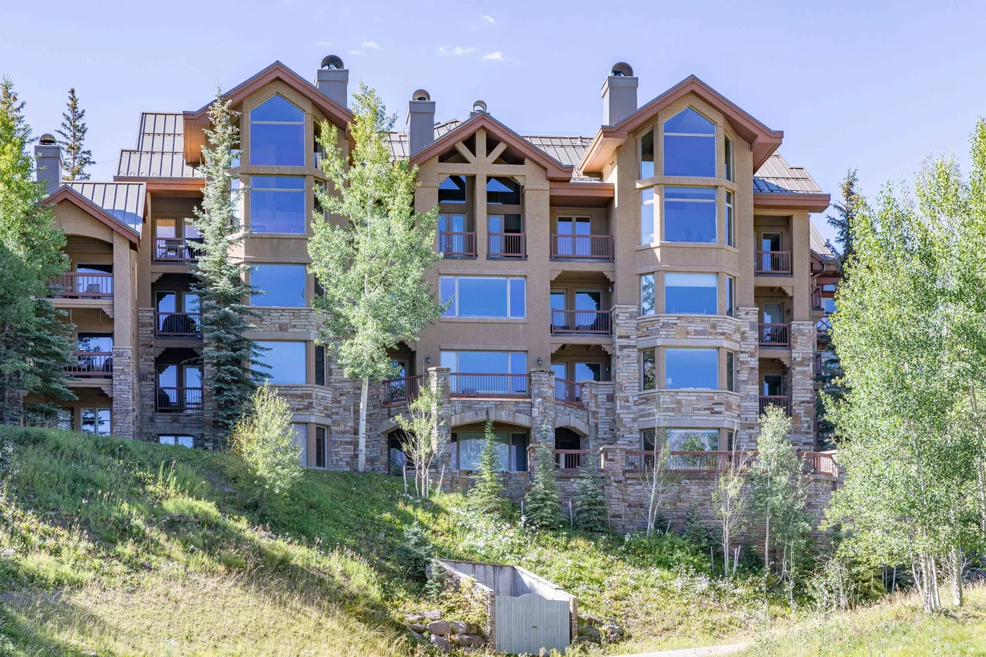 A multi-story building with stone walls, large windows, and trees on a grassy slope.