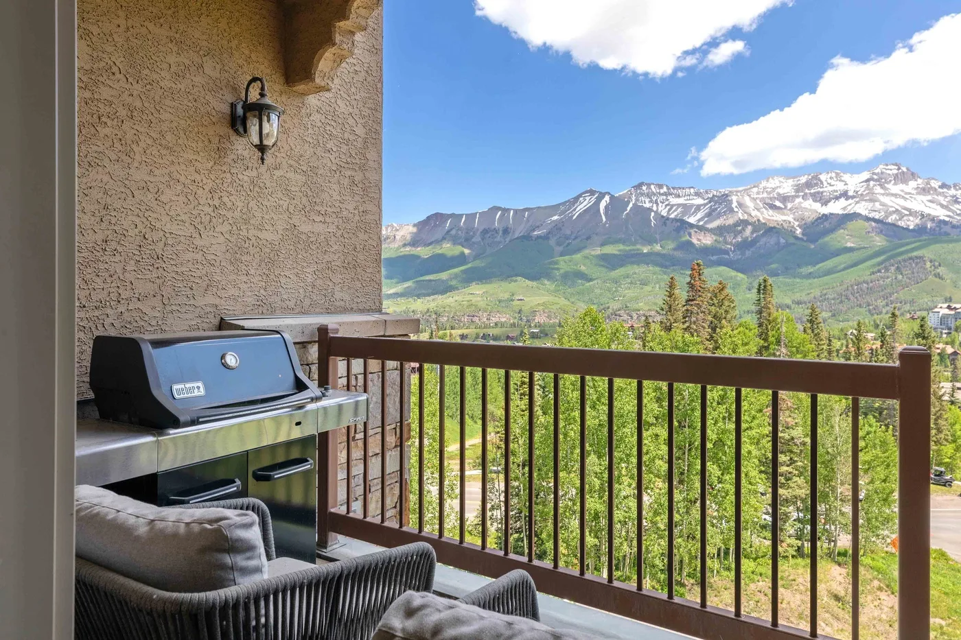 A balcony with seating and a grill overlooks a green valley and distant mountains.