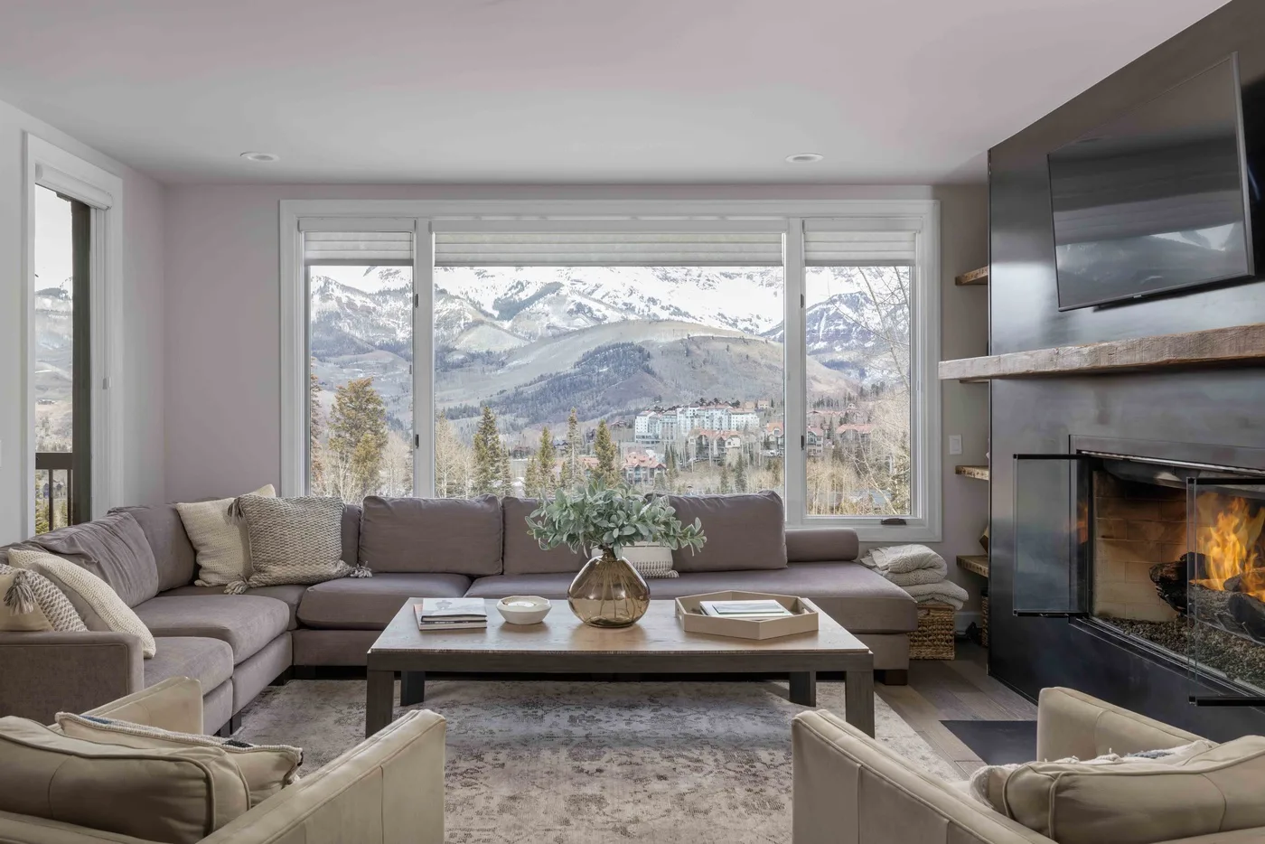 Una sala de estar con asientos alrededor de una mesa de centro, frente a grandes ventanas y montañas.