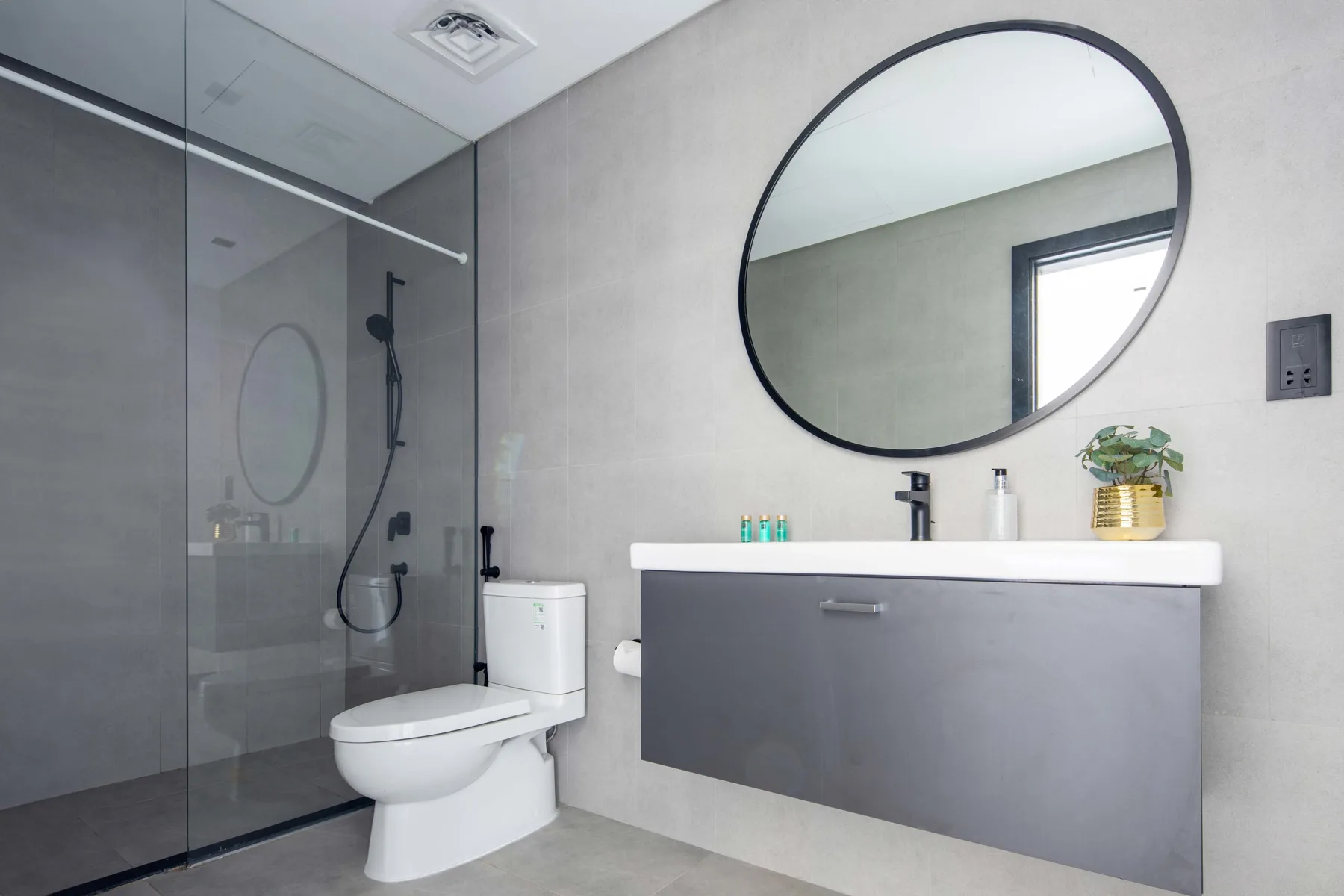 Un baño moderno con ducha de vidrio, inodoro y lavabo bajo un espejo redondo.