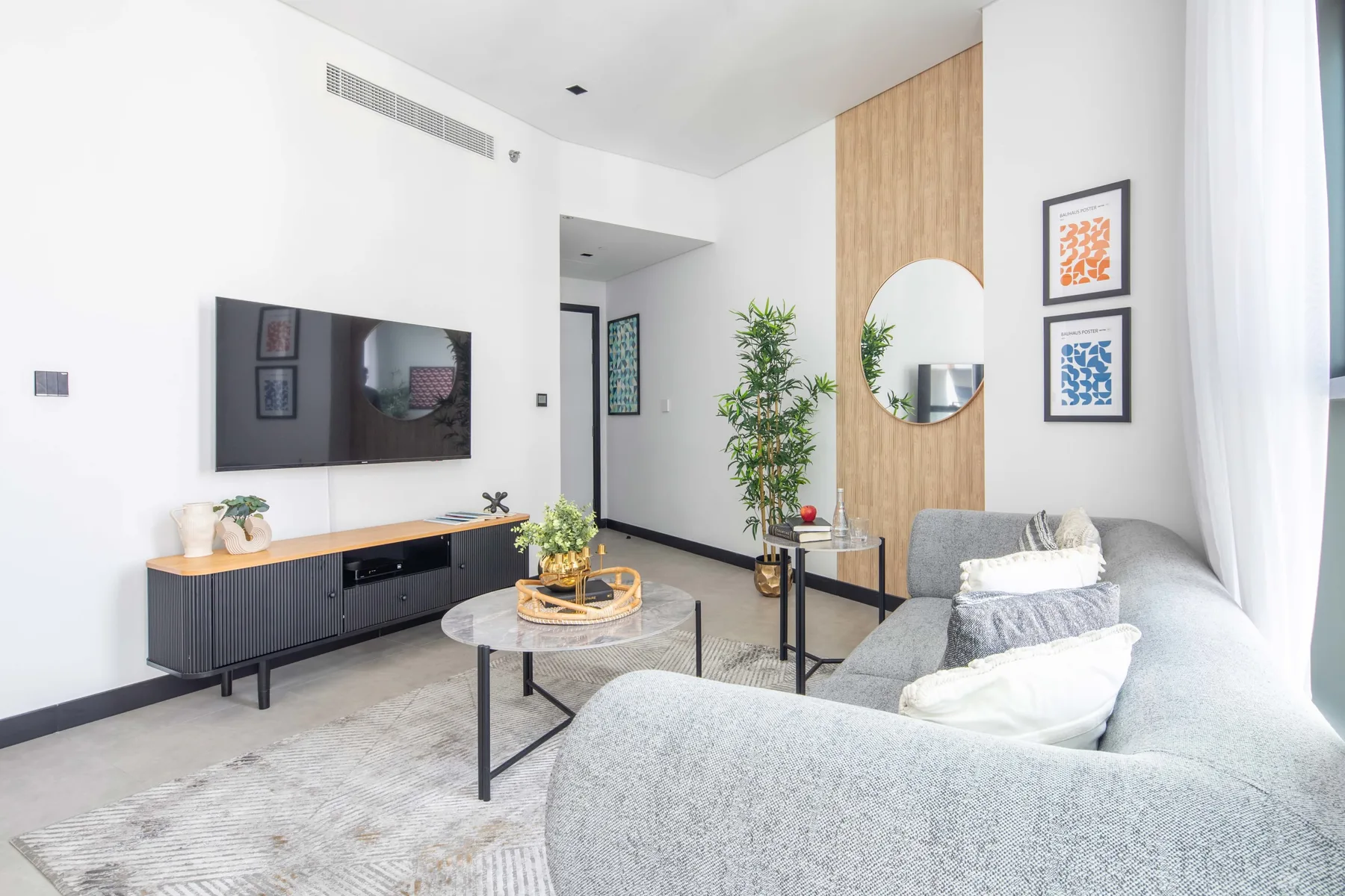 Una sala de estar luminosa con asientos grises, un televisor en la pared y una mesa de centro redonda.