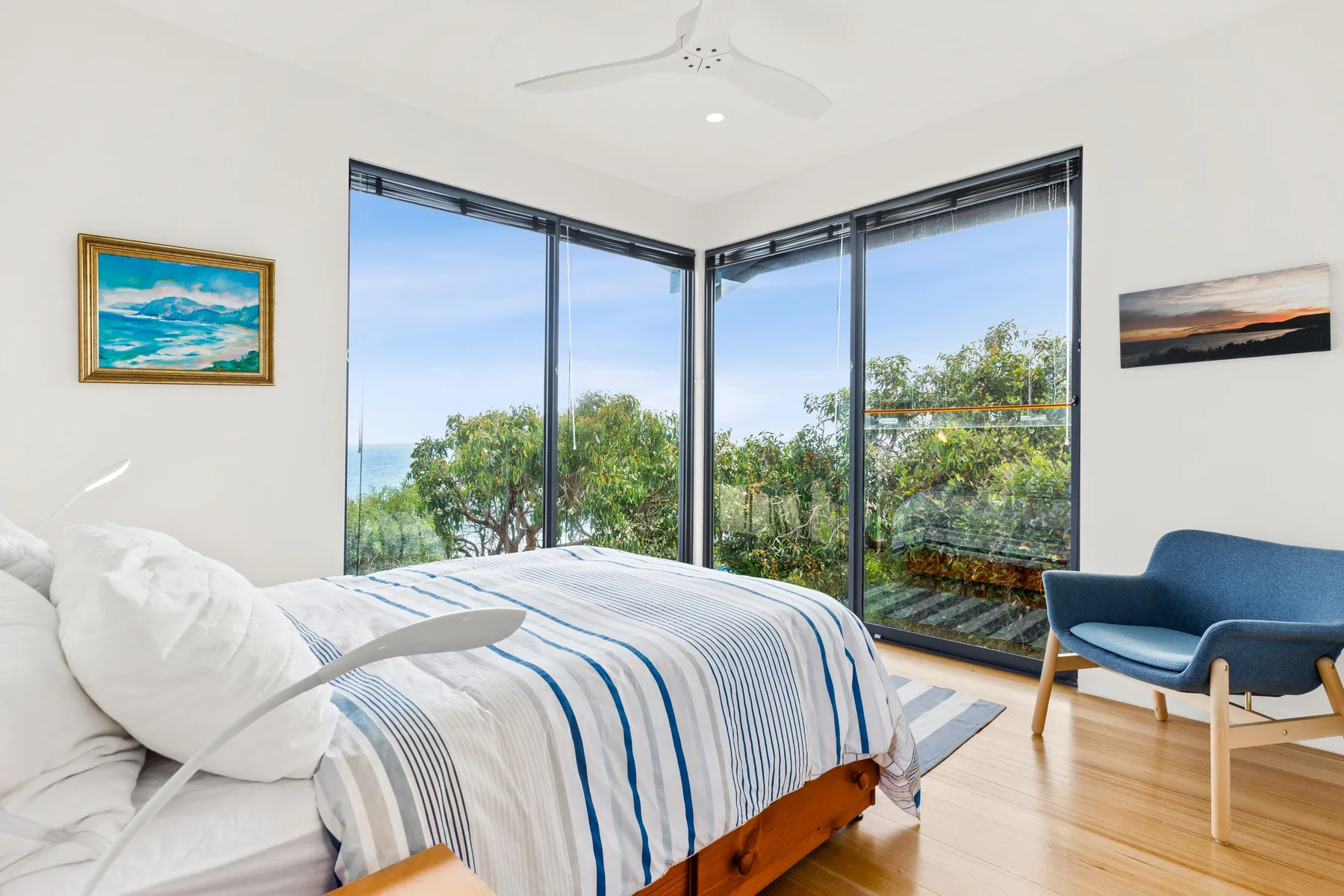 A bedroom with a bed beside large corner windows overlooking greenery.