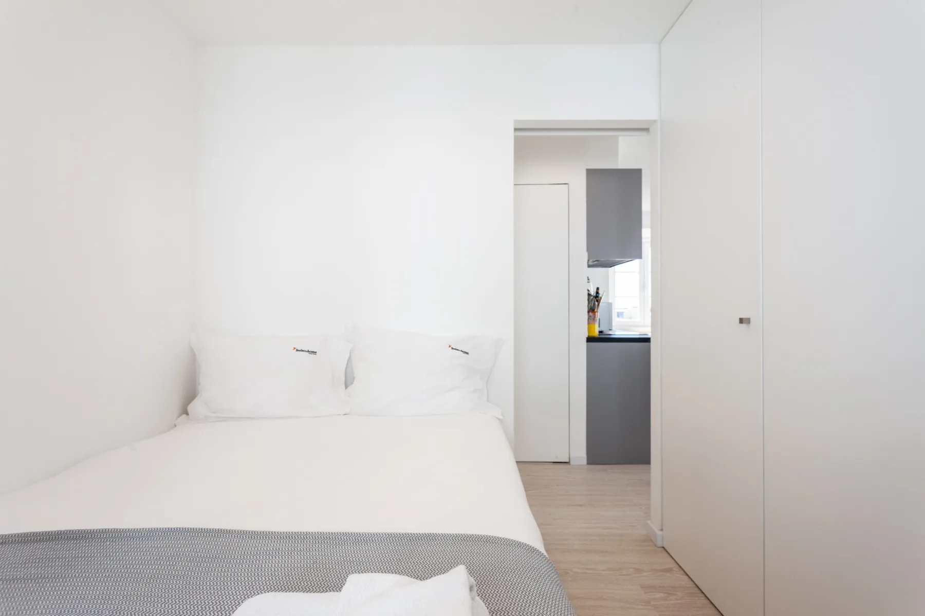 Un dormitorio luminoso con ropa de cama blanca y una puerta hacia la cocina.