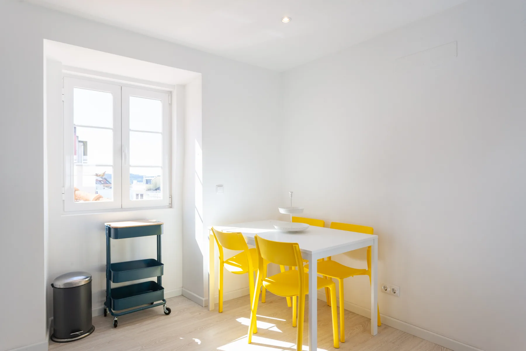 Comedor luminoso con una mesa blanca y asientos amarillos junto a una ventana.