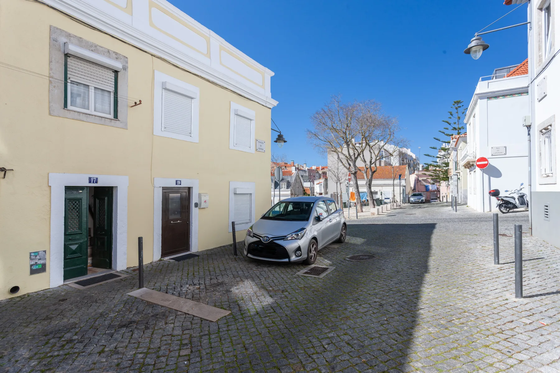 Una calle empedrada pasa entre edificios de colores claros, con un coche aparcado en primer plano.