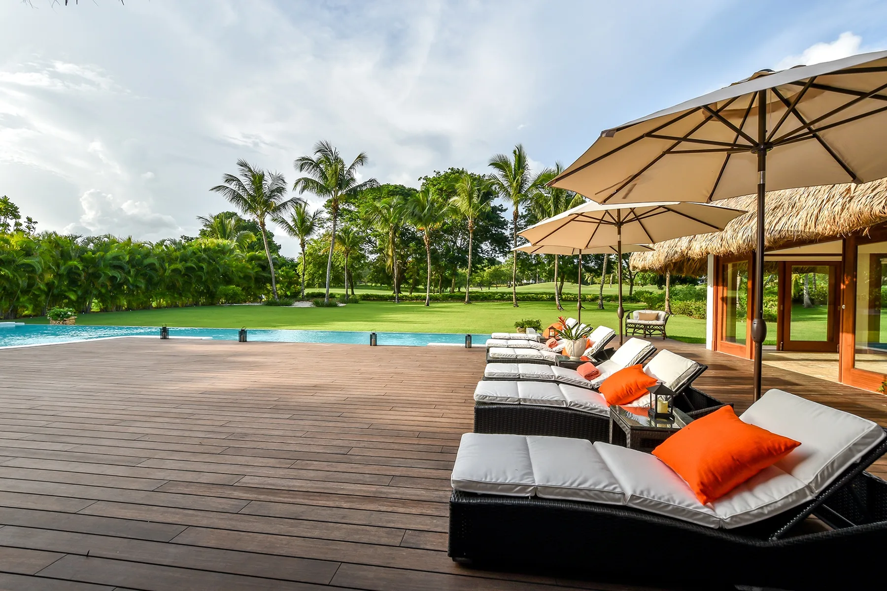Una terraza junto a la piscina tiene tumbonas acolchadas bajo grandes sombrillas.