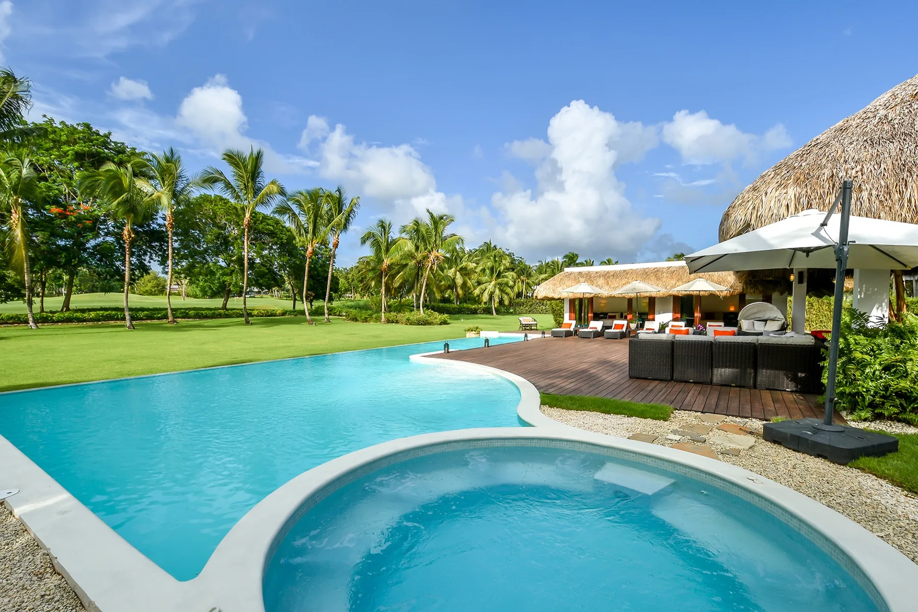 Una piscina exterior y un spa redondo están junto a una zona de descanso con techo de paja.