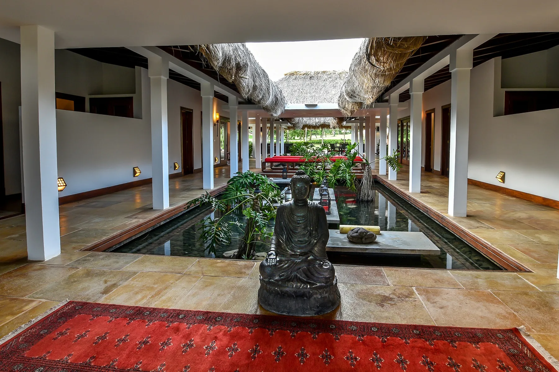 Un patio al aire libre con una larga piscina reflectante, plantas y una estatua sentada.