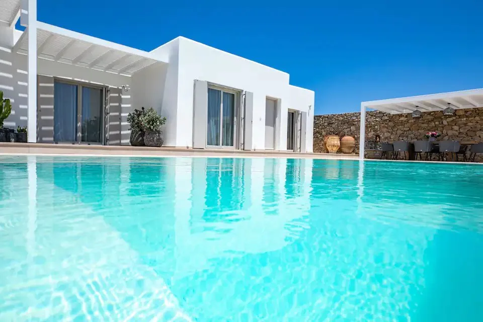 Una piscina junto a un edificio blanco y una zona de asientos al aire libre con sombra.