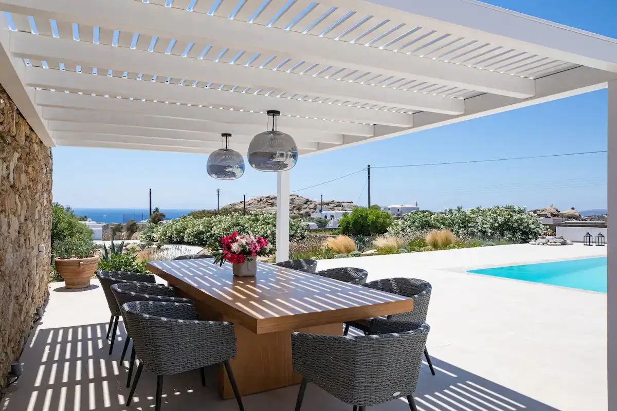 Una zona de comedor exterior a la sombra da a una piscina y al mar.