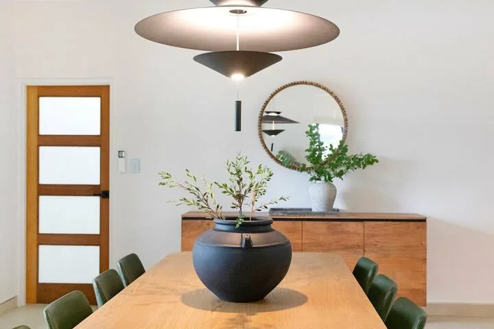 Un comedor con una mesa de madera, un centro decorativo oscuro y plantas frondosas.