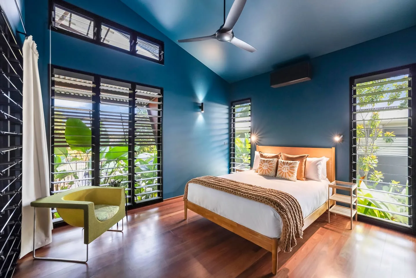 Dormitorio con una cama, paredes azules y grandes ventanas con vistas a la vegetación.