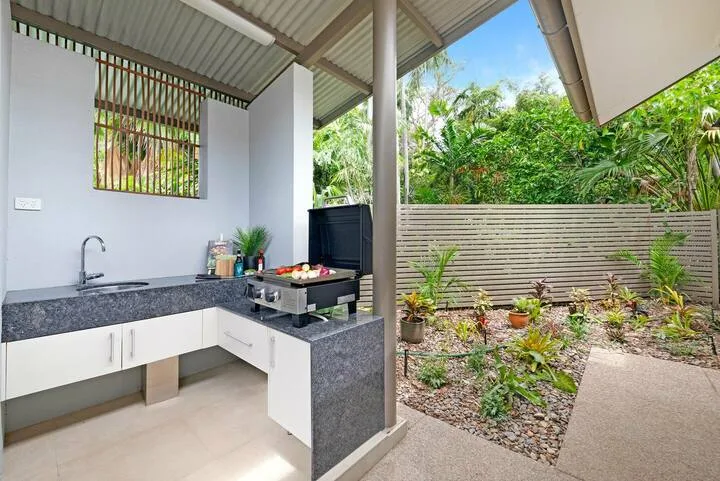 Una cocina exterior cubierta da a un jardín con plantas frondosas.