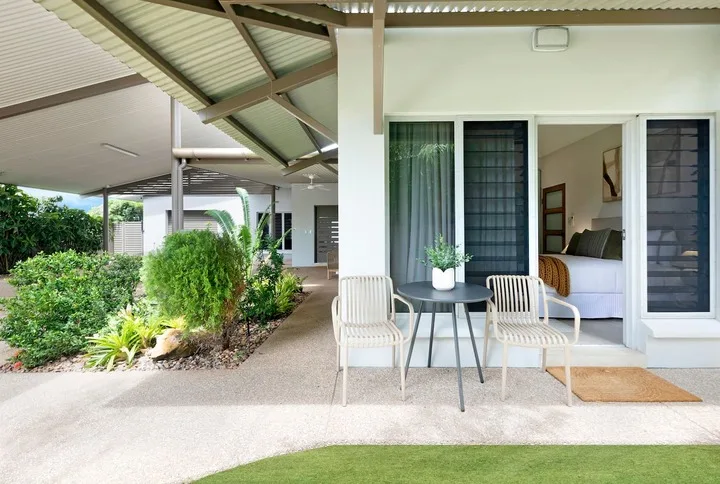 Un patio cubierto tiene una mesa pequeña y asientos junto a una habitación abierta.