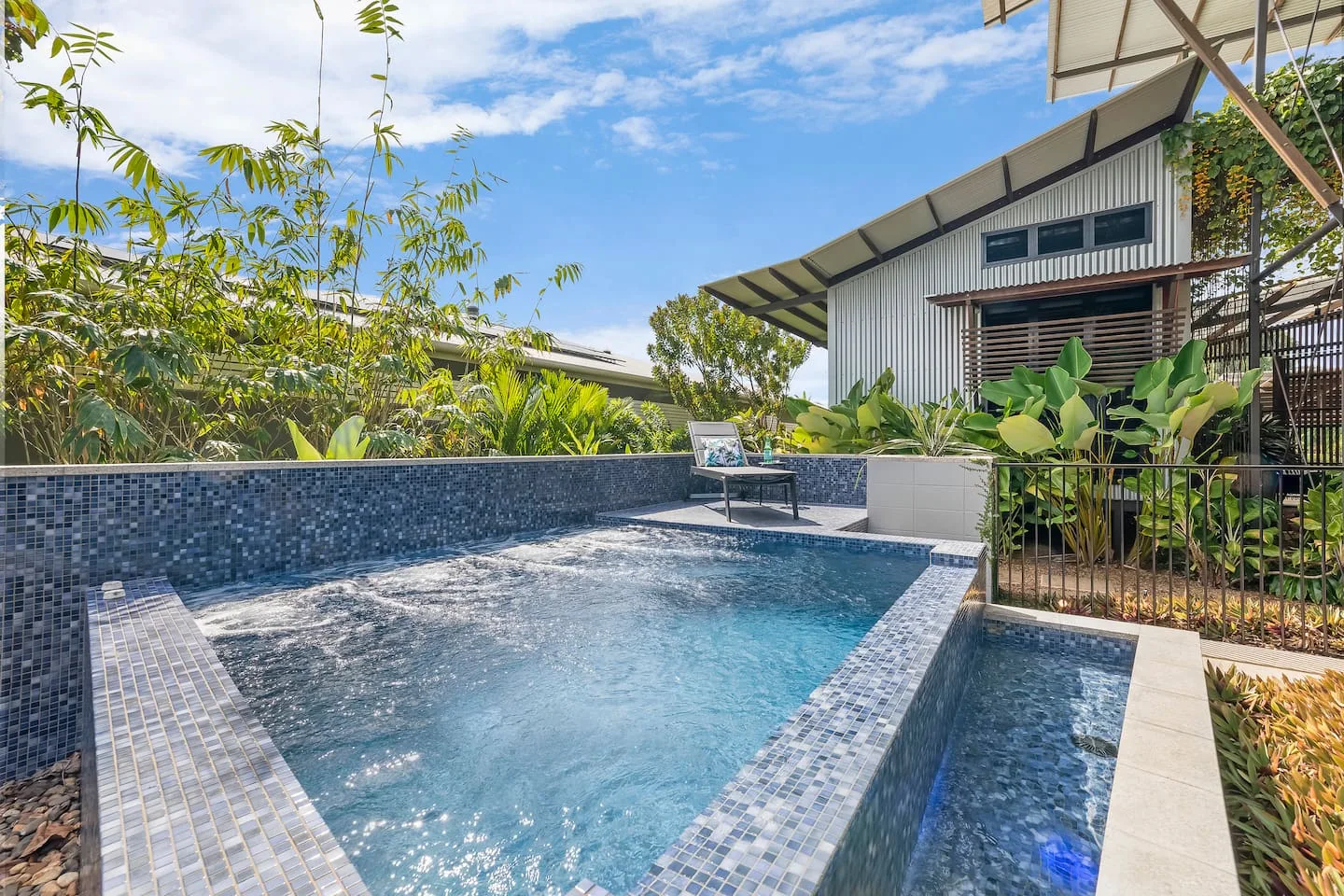 Una piscina al aire libre está junto a una casa y un jardín frondoso.