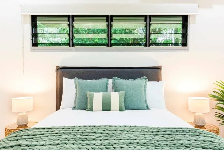 Una cama ordenada en un dormitorio, con ropa de cama y cojines verde azulado bajo una ventana horizontal.