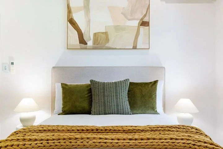 Un dormitorio con una cama bien tendida y almohadas bajo un cuadro abstracto.