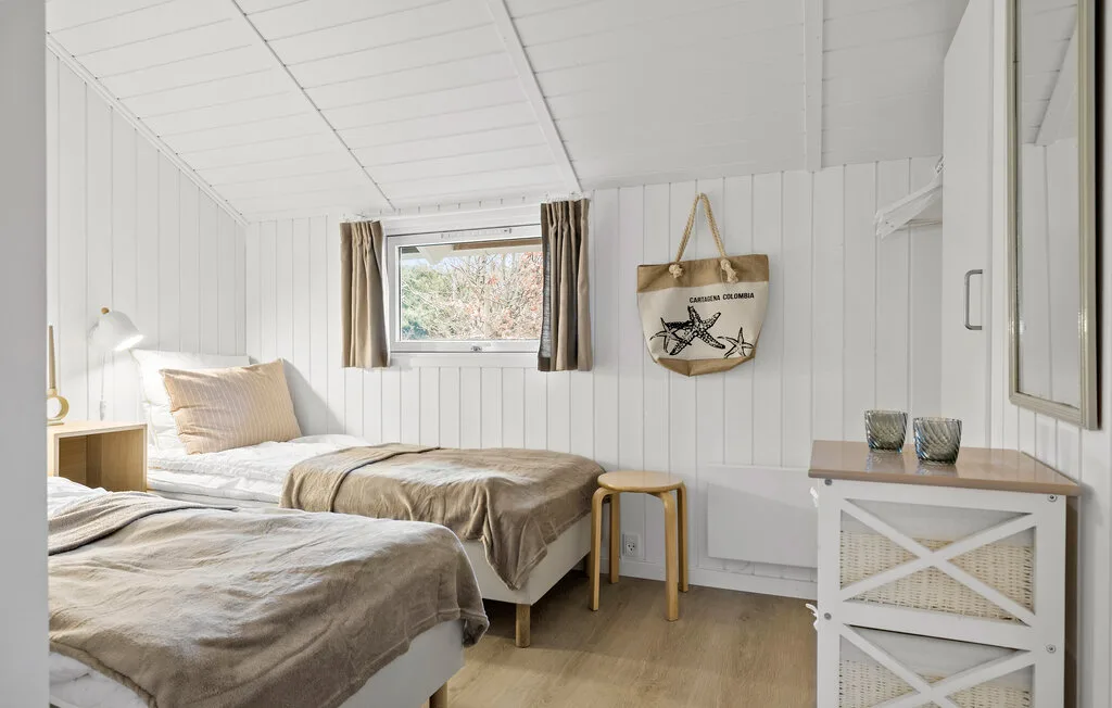 A bright bedroom with two beds, a small window, and white paneled walls.