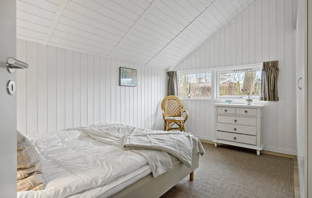 A bright bedroom with a bed, a wicker chair, and a window.