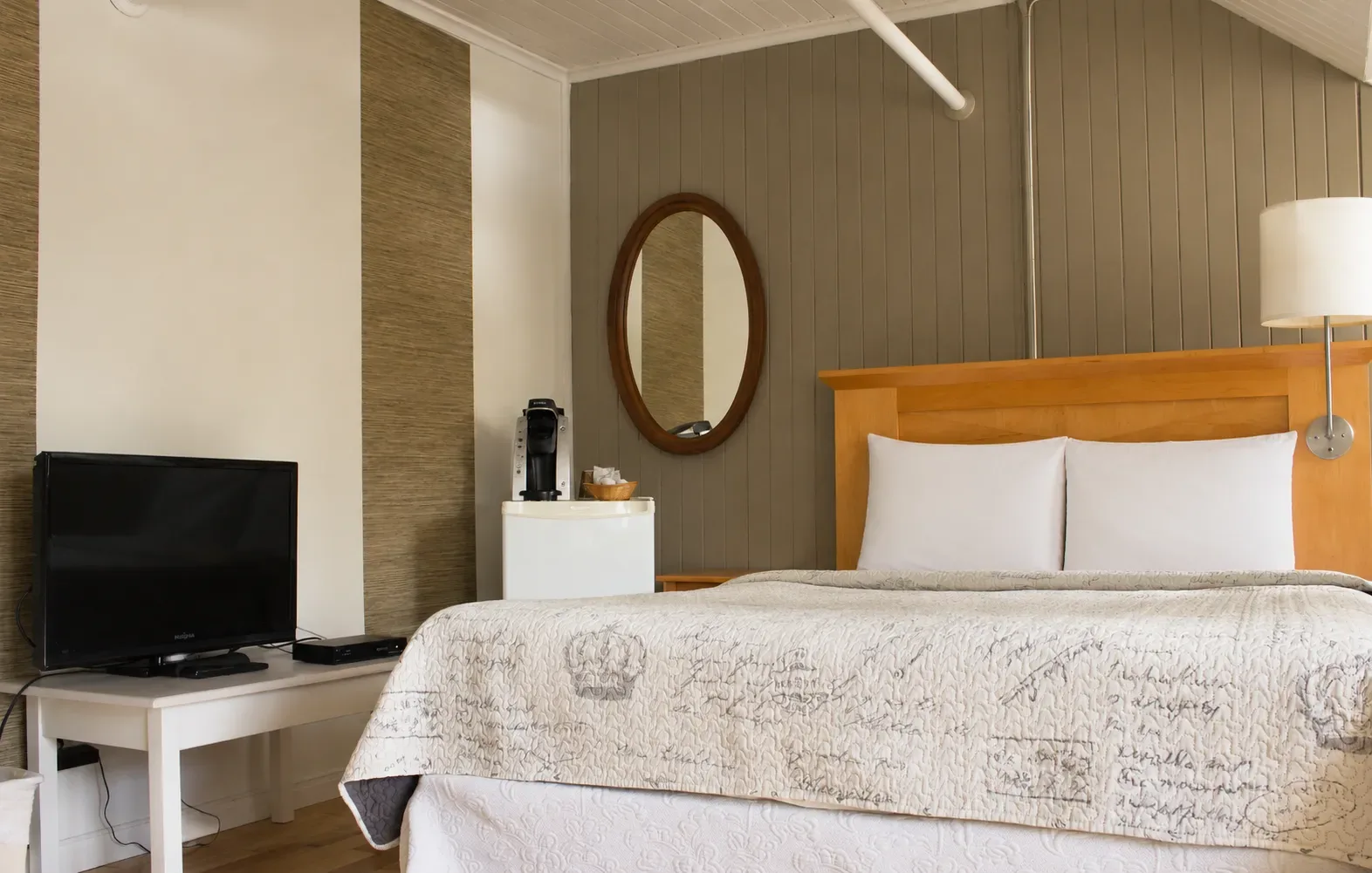 Bedroom with white bedspread, wooden dresser, chair, and lamp in a simple, bright room