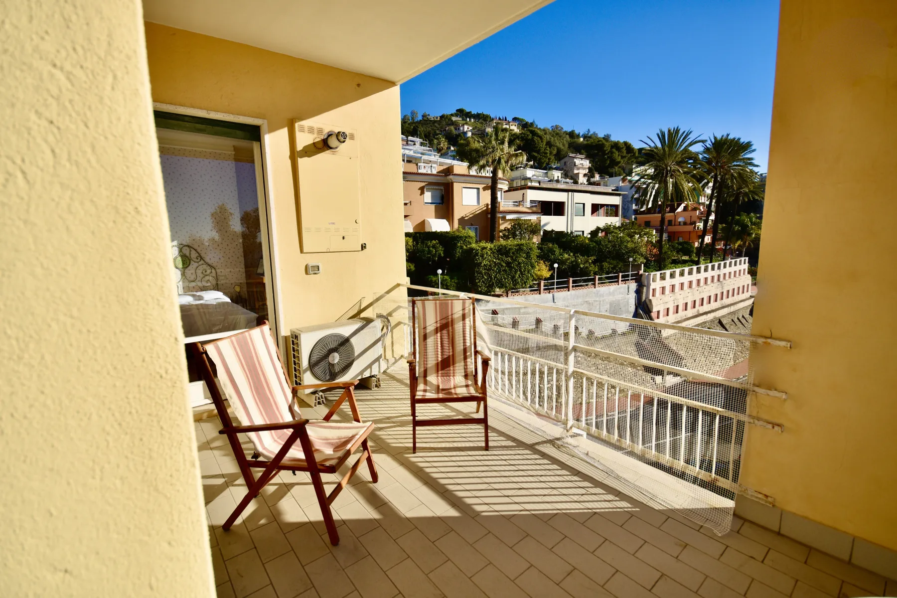 A sunny balcony with seating and a view of nearby buildings and greenery.