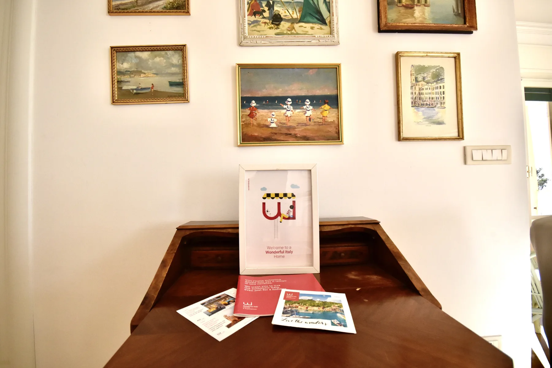 A wooden desk sits beneath a wall of framed paintings and a display print.