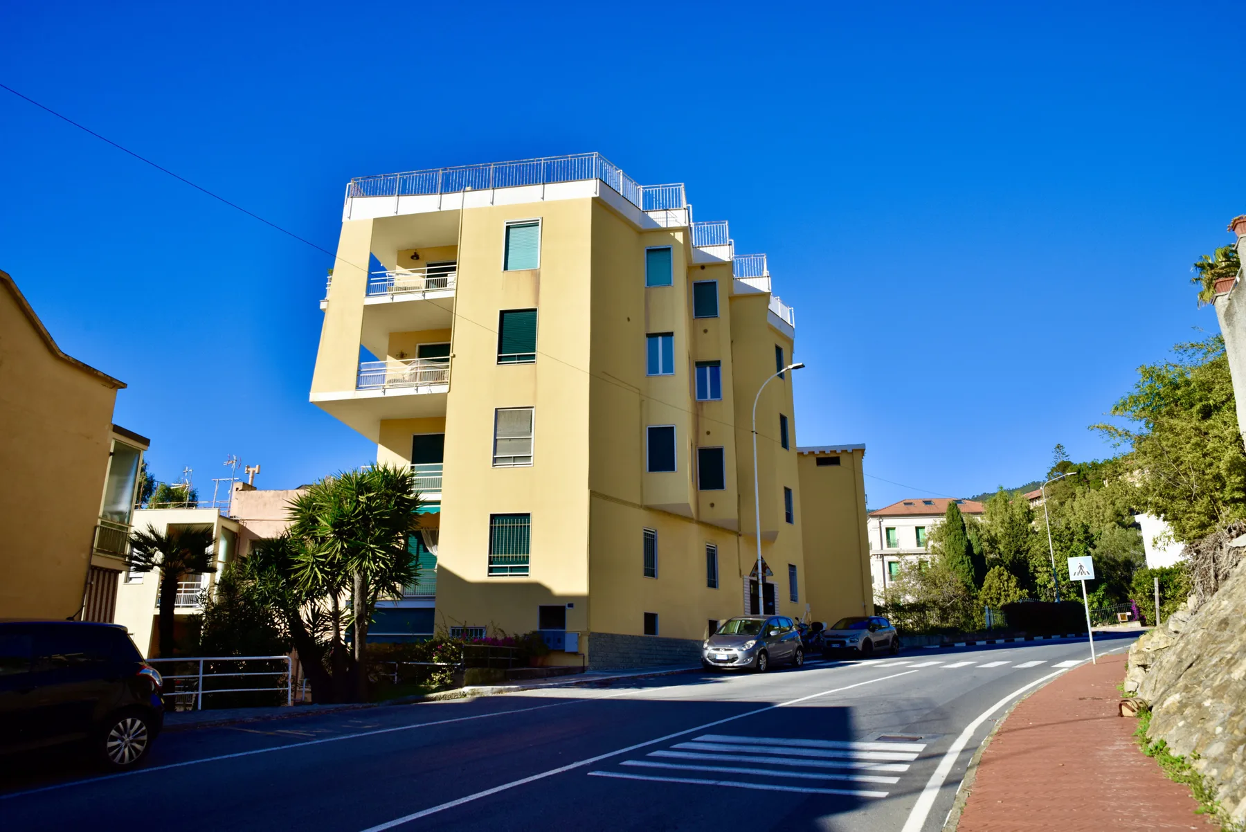 A yellow apartment building stands beside a curving road under a clear blue sky.